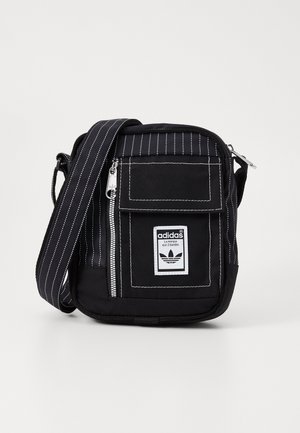 Sac bandoulière Adidas noir à fines rayures blanches, poche avant zippée, poche à rabat et patch logo de marque en blanc et noir.