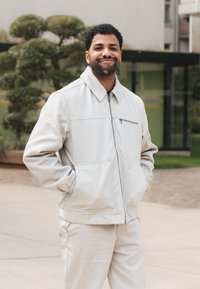 Jasnoszara kurtka zapinana na zamek z kołnierzykiem, kieszenią na zamek i przednimi kieszeniami, w połączeniu z pasującymi jasnoszarymi spodniami o gładkim, teksturowanym materiale.