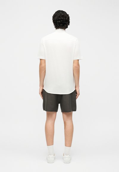 Νεαρός που στέκεται με την πλάτη γυρισμένη, φοράει λευκό κοντομάνικο πουκάμισο, σκούρα σορτς, άσπρες κάλτσες και άσπρα αθλητικά παπούτσια σε απλό φόντο.