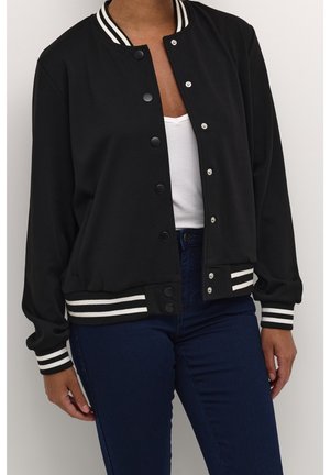 Person iført sort varsityjakke med hvide striber på ærmer og linning, hvid top og mørkeblå jeans, stående mod hvid baggrund.