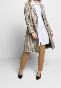 Randig trenchcoat i gult, svart och vitt; enkelknäppt med stora knappar. Bärs över en vit skjorta och tan-färgade, åtsittande byxor. Svarta skor.