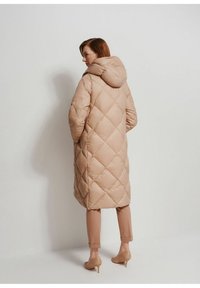 Puchowa zimowa kurtka w jasnym beżu z pikowanym wzorem w diamenty, z kapturem i w pełnej długości. Połączona z dopasowanymi beżowymi spodniami i szpilkami.