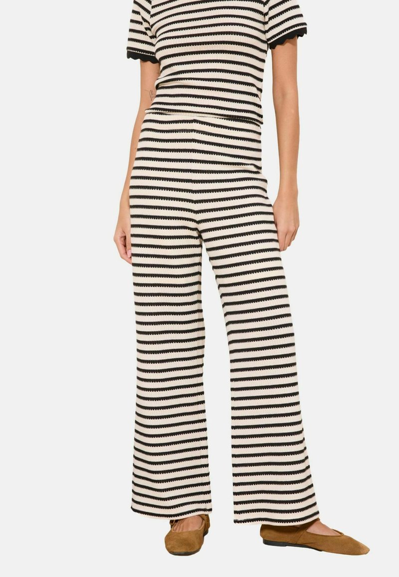 Femme portant un pantalon en maille à rayures horizontales noires et blanches et un haut manches courtes assorti avec des chaussures plates marron.