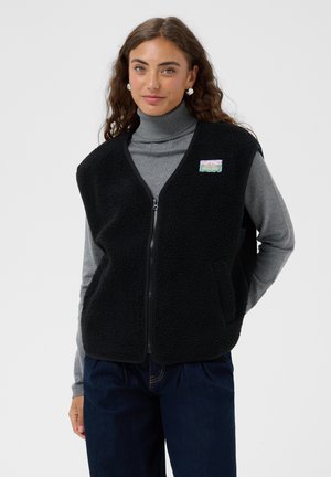 Femme portant une veste polaire noire zippée par-dessus un col roulé gris et un pantalon bleu foncé, debout avec une main derrière le dos.