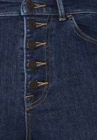 Vaqueros de cintura alta en azul oscuro con cinco botones de acabado negro y costuras en dorado en contraste a lo largo de la costura.