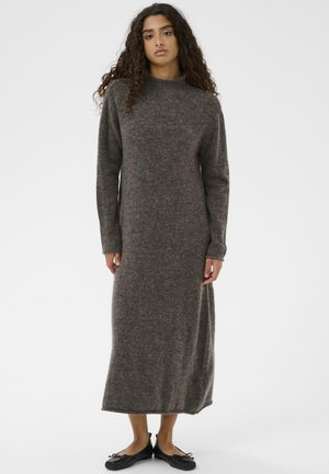 Junge Frau mit langen, lockigen Haaren, die ein graues Strickkleid mit langen Ärmeln und Knöchellänge sowie schwarze Ballettschuhe vor einem weißen Hintergrund trägt.