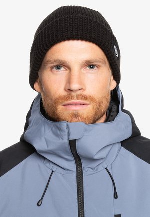Blå og sort jakke med højt krave og lynlås, parret med en ribbet sort beanie. Har en struktureret stof.