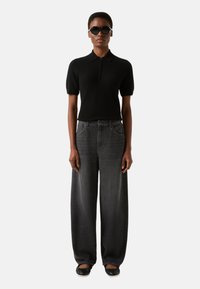 Czarna koszulka polo z krótkim rękawem i zamkiem błyskawicznym, zestawiona z szerokimi szarymi dżinsami. Dodatki to okrągłe, ciemne okulary przeciwsłoneczne i czarne płaskie buty.