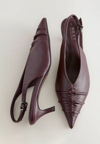 Burgundské kožené slingback topánky s špicatou špičkou, zhrnutým predným detailom a stredne vysokým opätkom. Nastaviteľný pásik okolo členka so sponou.