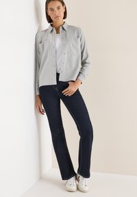 Szara koszula z guzikami, z dwoma przednimi kieszeniami, noszona na białym T-shircie, w połączeniu z ciemnoniebieskimi dżinsami w stylu flare i białymi trampkami.