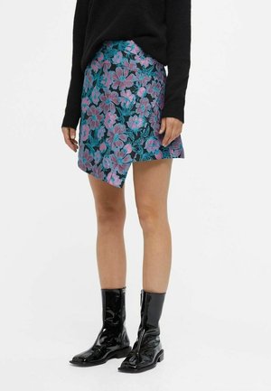Frau trägt schwarzen Pullover, blau-pink geblümten asymmetrischen Rock und glänzende schwarze Stiefel bis zur Mitte der Wade, steht vor einem einfarbigen Hintergrund.