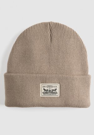 Brown knitted beanie featuring a folded cuff and a rectangular label with black text and graphic. Textured ribbed fabric.