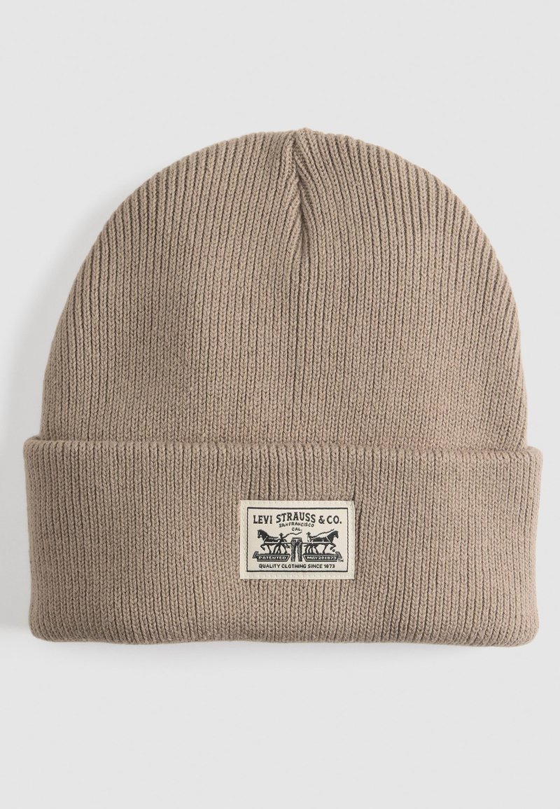 Bruine gebreide beanie met een omgeslagen rand en een rechthoekig label met zwarte tekst en grafiek. Gestructureerde ribbelstof.