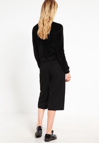 Donna con capelli biondi lunghi, che indossa un top nero a maniche lunghe, pantaloni neri cropped a gamba larga e scarpe oxford nere, in piedi su un pavimento bianco.