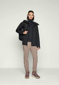 Chaqueta negra impermeable con cremallera, superpuesta sobre una camiseta negra de manga larga. Pantalones beige y botas de senderismo marrones. Lleva una mochila.