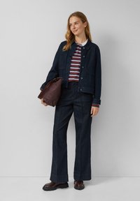 Vrouw in een donkere spijkerjas en broek, gestreept poloshirt, met een grote donkerbruine leren tas, staande tegen een effen achtergrond.