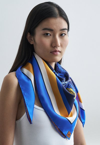 Sieviete ar gariem tumšiem matiem valkā baltu bezpiedurkņu t-kreklu un lielu zilu, dzeltenu un balto rakstainu zīda šalli ap kaklu uz vienkārša fona.