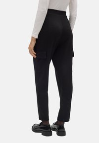 Pantalones cargo negros a medida con piernas ajustadas, que cuentan con bolsillos laterales y una textura suave, combinados con unos zapatos negros brillantes.