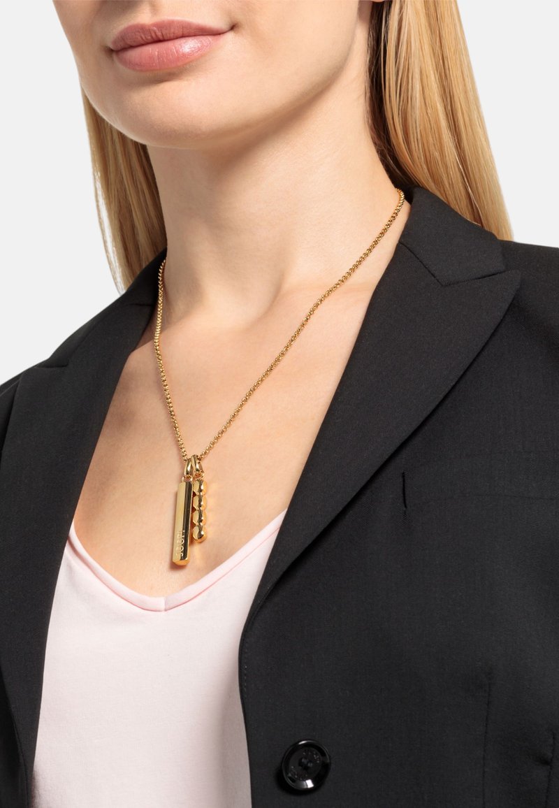Gouden ketting met een rechthoekige hanger en een parelaccent, hangend aan een dunne ketting, gedragen over een roze top en een zwart blazertje.