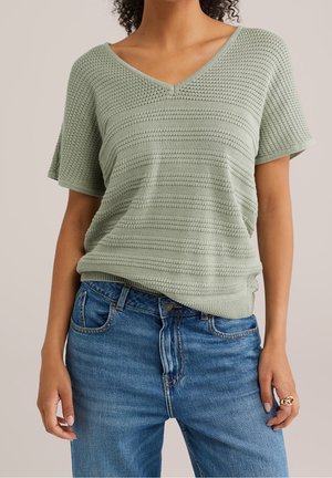 Kobieta ubrana w jasnorozowy teksturowany krótkim rękawem sweter z dekoltem w serek i niebieskie dżinsy, pokazująca tułów i ręce.