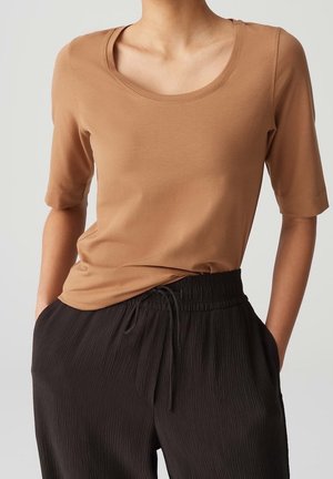 Personne portant un t-shirt marron à manches courtes et col rond, rentré dans un pantalon noir texturé à cordon, les mains dans les poches.