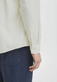 Camisa de lino color crema con puños abotonados, tela texturizada y dobladillo desaliñado, combinada con pantalones de un oscuro color navy que presentan un bolsillo trasero.