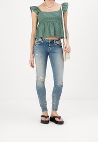 Top verde, ajustado, de manga corta con acentos de volantes, combinado con unos pantalones vaqueros ajustados, desgastados, de color azul claro y chanclas negras.