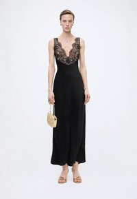 Γυναίκα που φοράει ένα μακρύ μαύρο φόρεμα με δαντελένιο λαιμόκοψη, κρατώντας μια μικρή μπεζ τσάντα και φορώντας μπεζ πέδιλα με ανοιχτά δάχτυλα σε λευκό φόντο.