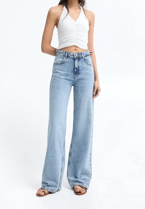 Kobieta ubrana w jasnoniebieskie jeansy z wysokim stanem i szerokimi nogawkami, biały marszczony crop top na ramiączkach i brązowe sandały z odsłoniętymi palcami na jednolitym białym tle.