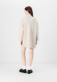 Beige, oversized strikket kjole med høy krage, lange ermer og sidesplitter, båret med svarte sko og hvite sokker.