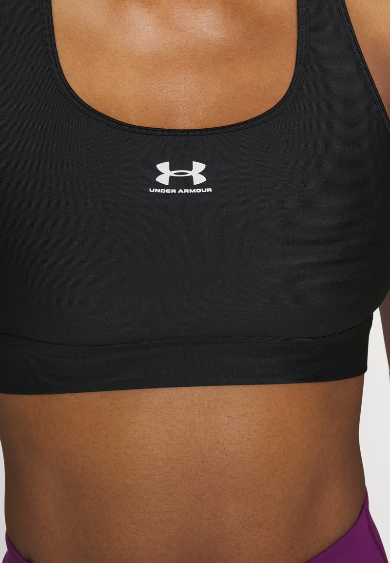 Sujetador deportivo negro hecho de una tela con textura suave, que presenta un logo blanco de Under Armour en la zona del pecho.