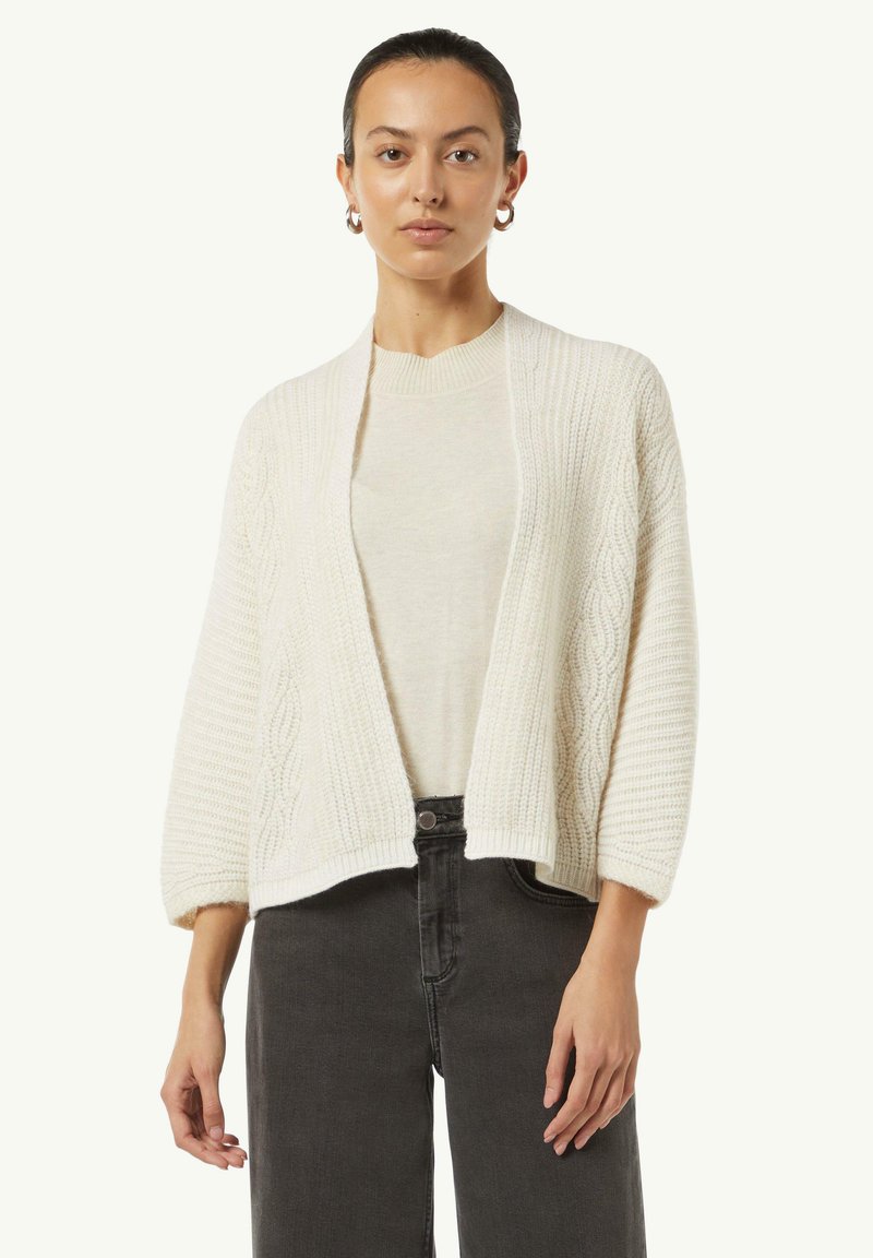 Kremowy, dzianinowy kardigan z wzorem w warkocze, otwarty z przodu, z rękawami o długości trzech czwartych, połączony z lekkim swetrem i ciemnymi dżinsami.