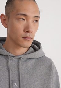 La sudadera gris presenta un parche con el logo, cordones y una textura acanalada. La tela parece suave, con un ajuste relajado y un diseño clásico con capucha.
