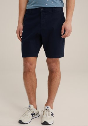 Mand iført marineblå shorts, lyseblå mønstret skjorte og hvide sneakers med marineblå detaljer, stående på en neutral baggrund.