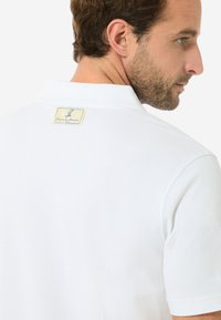 Das weiße Polo-Shirt hat einen klassischen Kragen und kurze Ärmel, mit einem etiket am hinteren Hals, das die Markendetails präsentiert.