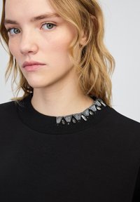 Czarna bluza z ribbowym kołnierzem, zdobiona metalowymi i szklanymi detali ułożonymi w trójkątny wzór.