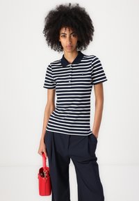 Stripet marineblå og hvit polo med krage og fem knapper, paret med løse marineblå bukser og en liten rød håndveske.