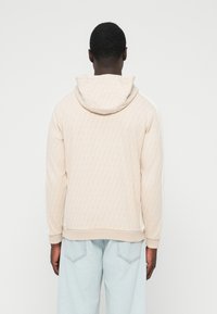 Svetlo bež majica s kapuco s teksturiranim vzorcem diamantov, opremljena z zategovalnim vrvico na kapuci in belimi stranskimi poudarki, združena s svetlo modrimi jeans hlačami.