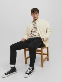 Light beige long-sleeve shirt over a striped brown and white t-shirt, paired with black pants and black high-top sneakers. Simple wooden chair.