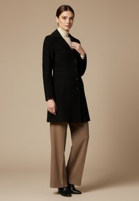 Vrouw die een zwarte jas met knopen draagt, beige wijd uitlopende broek, zwarte enkellaarzen en een witte coltrui tegen een beige achtergrond.