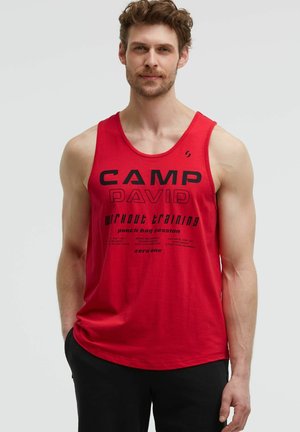 Mann trägt ein rotes Tanktop mit schwarzem Workout-Trainingstext, steht mit einer Hand in der Tasche vor einem einfarbigen Hintergrund.