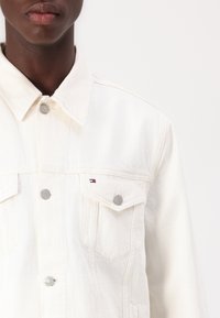 Jachetă din denim alb cu guler ascuțit, două buzunare pe piept și nasturi gri. Prezintă o mică emblema în roșu, alb și albastru.