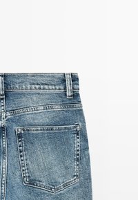 Zadní kapsa a poutko na pásek modrých džínů s viditelným šitím na bílém pozadí.