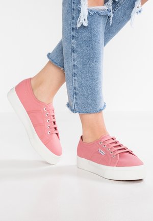 Personne portant un jean bleu clair déchiré et des baskets compensées en toile rose avec des semelles blanches, debout sur un sol blanc.