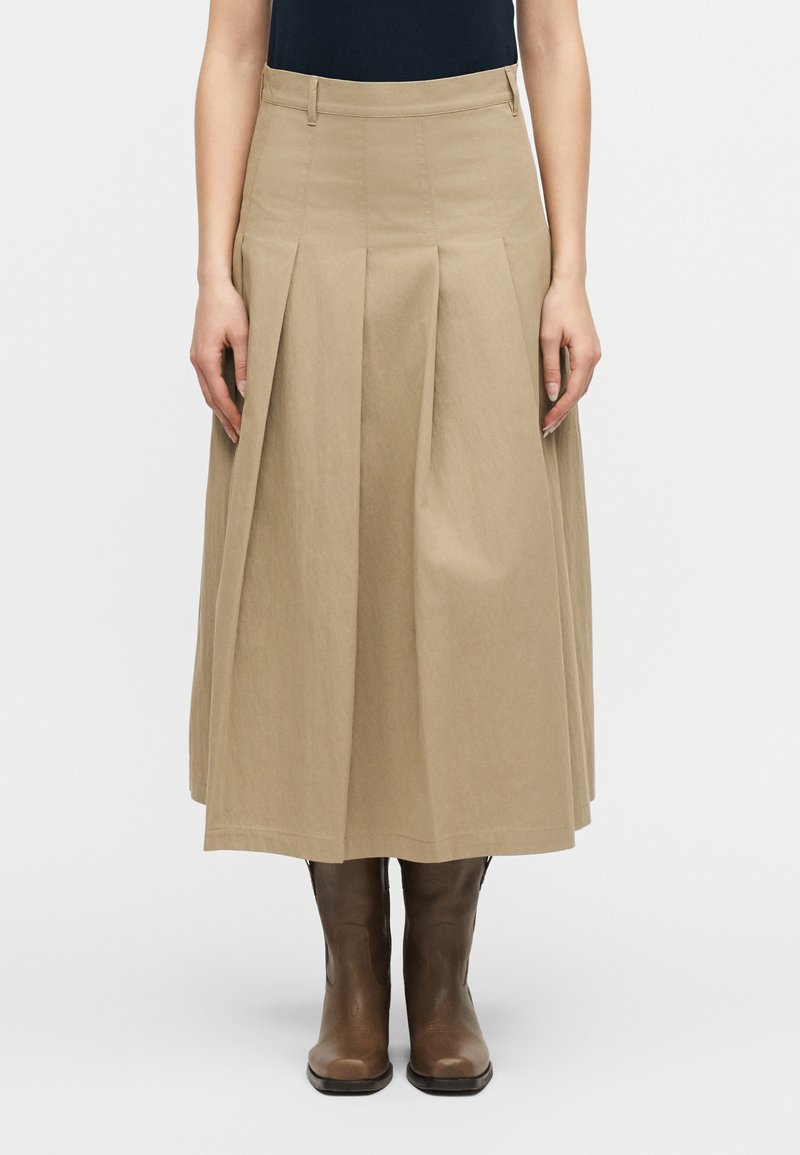 Γυναίκα που φοράει μπεζ πλισέ midi φούστα συνδυασμένη με καφέ δερμάτινα μποτάκια μέχρι τον αστράγαλο, στέκεται μπροστά σε απλό λευκό φόντο.