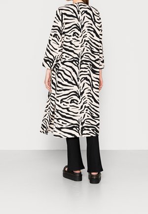 Femme portant un long manteau à rayures zèbre avec de larges manches sur un pantalon noir et des sandales compensées, debout devant un fond uni.