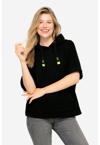 Felpa nera trapuntata con maniche corte e tasca a marsupio, con dettagli e stringhe neon verdi. Vengono indossati jeans grigi.