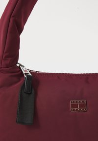 Geantă de umăr din nylon burgund cu o curea texturată neagră, fermoar argintiu și un accent metalic pătrată cu marcaj pe partea din față.