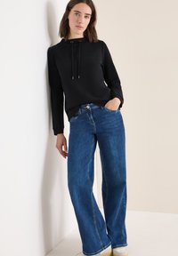 Schwarzer Pullover mit Kapuze und Kordelzug, kombiniert mit weit geschnittenen blauen Jeans mit Fronttaschen und sichtbaren Nähten.