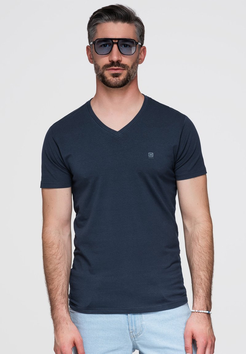 Homme aux cheveux courts et barbe portant de grandes lunettes de soleil foncées, un T-shirt bleu marine à col en V et un jean bleu clair, debout devant un fond uni.